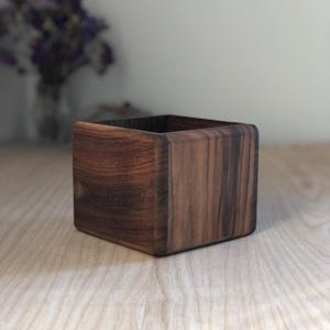 Puede incluir: Una caja de madera cuadrada hecha de madera de nogal oscuro. La caja tiene un acabado liso y pulido y es perfecta para guardar objetos pequeños.