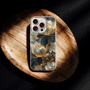 Puede incluir: Funda transparente para tel&eacute;fono con un dise&ntilde;o floral con flores azules, doradas y blancas. La funda est&aacute; sobre una bandeja de madera con un fondo oscuro. El estampado floral tiene un aspecto art&iacute;stico detallado.