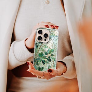 Puede incluir: Una funda de tel&eacute;fono inteligente con un dise&ntilde;o bot&aacute;nico. La funda presenta un patr&oacute;n de hojas verdes y detalles dorados sobre un fondo claro. La funda tiene un recorte para la c&aacute;mara y la sostiene una persona.