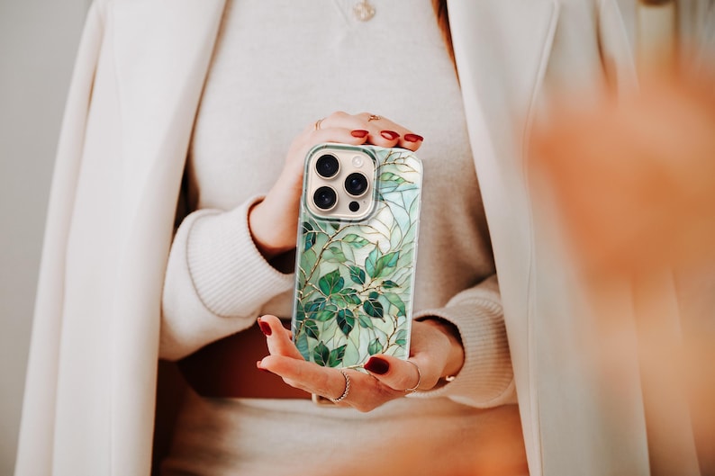 Puede incluir: Una funda de tel&eacute;fono inteligente con un dise&ntilde;o bot&aacute;nico. La funda presenta un patr&oacute;n de hojas verdes y detalles dorados sobre un fondo claro. La funda tiene un recorte para la c&aacute;mara y la sostiene una persona.