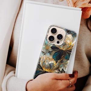 Puede incluir: Una funda transparente para tel&eacute;fono con un dise&ntilde;o floral en tonos dorados, verde azulado y blanco, sujeta contra un libro blanco. La funda presenta un patr&oacute;n art&iacute;stico detallado con contornos oscuros. La funda del tel&eacute;fono la sostiene una persona que lleva un su&eacute;ter y una blazer color crema.