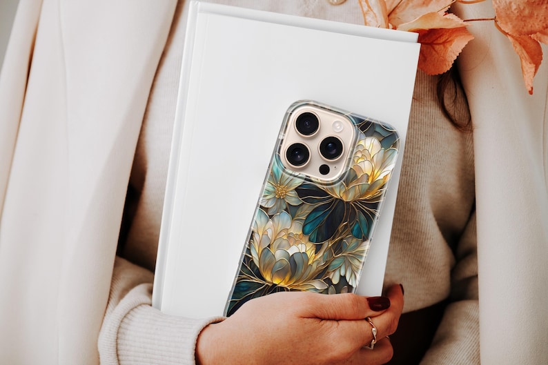 Puede incluir: Una funda transparente para tel&eacute;fono con un dise&ntilde;o floral en tonos dorados, verde azulado y blanco, sujeta contra un libro blanco. La funda presenta un patr&oacute;n art&iacute;stico detallado con contornos oscuros. La funda del tel&eacute;fono la sostiene una persona que lleva un su&eacute;ter y una blazer color crema.