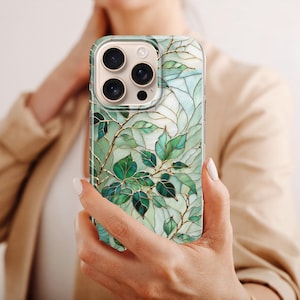Puede incluir: Funda de tel&eacute;fono inteligente con un dise&ntilde;o bot&aacute;nico. La funda presenta hojas verdes y vetas doradas sobre un fondo azul claro. La funda tiene un acabado brillante y un parachoques protector. La funda del tel&eacute;fono est&aacute; en la mano de una persona.