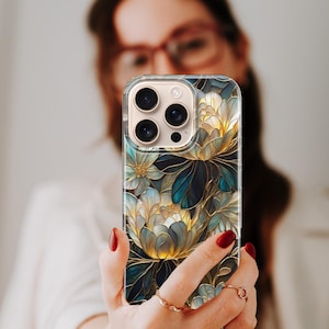 Puede incluir: Funda transparente para tel&eacute;fono con un dise&ntilde;o floral en tonos azules, dorados y blancos. La funda presenta una ilustraci&oacute;n detallada de flores y hojas estilizadas. La funda del tel&eacute;fono se sostiene frente a una persona.