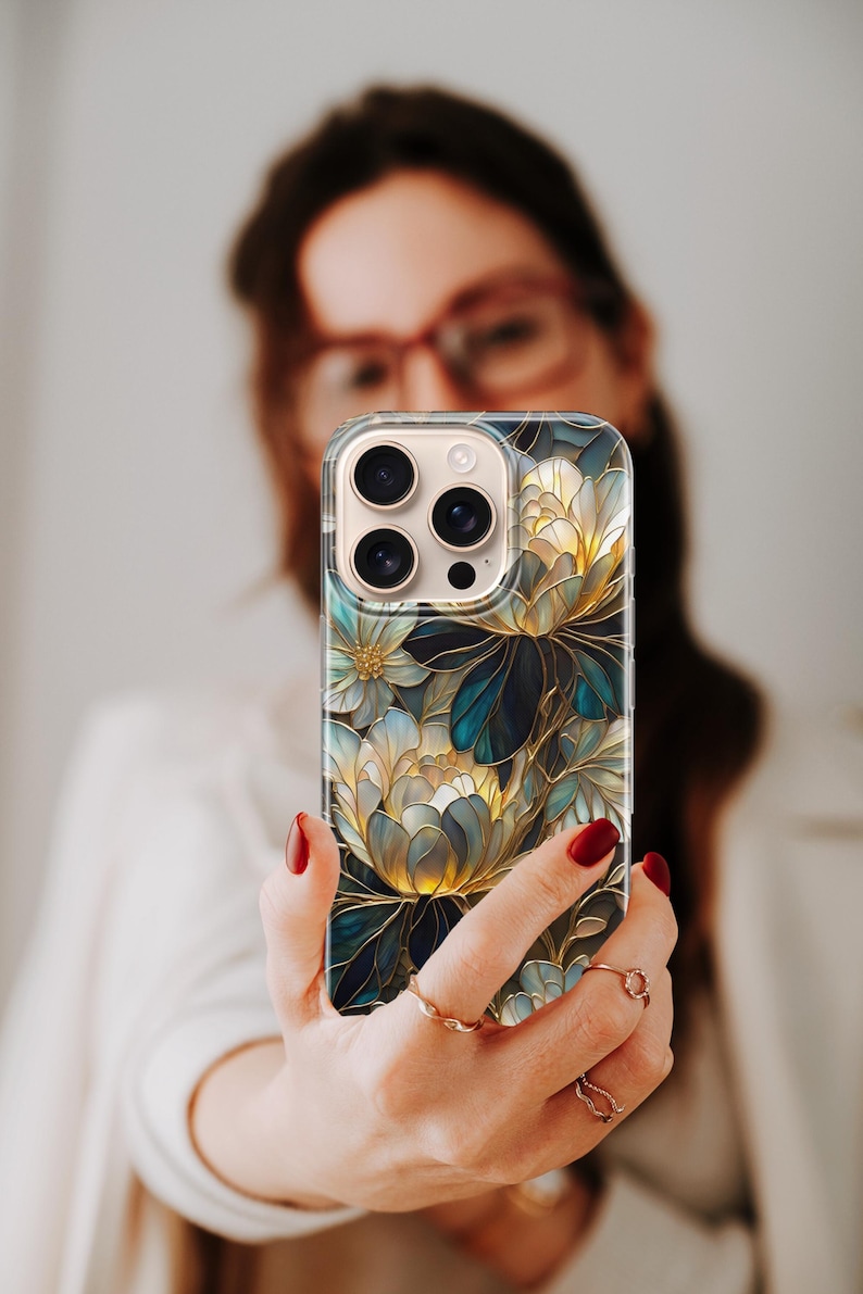 Puede incluir: Funda transparente para tel&eacute;fono con un dise&ntilde;o floral en tonos azules, dorados y blancos. La funda presenta una ilustraci&oacute;n detallada de flores y hojas estilizadas. La funda del tel&eacute;fono se sostiene frente a una persona.