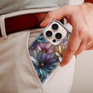 Puede incluir: Una funda transparente para tel&eacute;fono con un dise&ntilde;o floral con flores moradas, azules y amarillas. La funda es parcialmente visible en un bolsillo, con un cintur&oacute;n de cuero marr&oacute;n y un reloj con correa de cuero marr&oacute;n en el fondo.