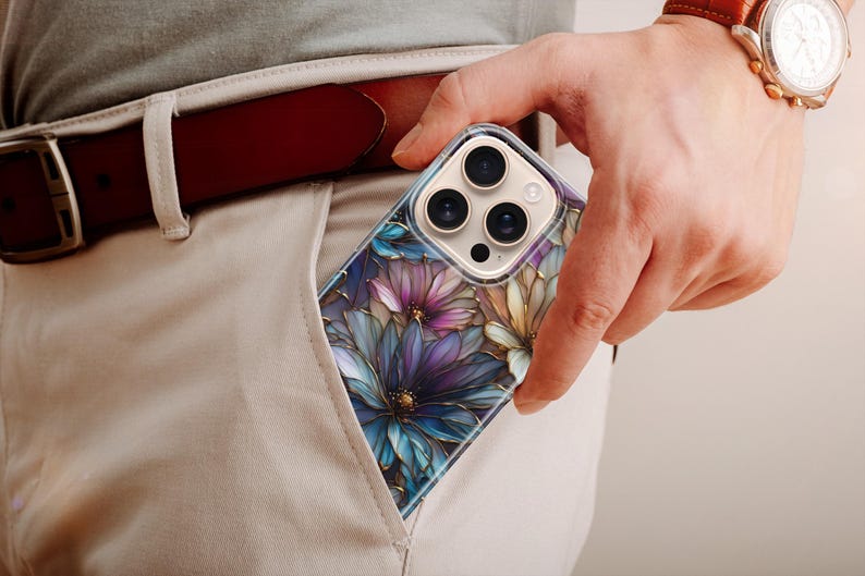 Puede incluir: Una funda transparente para tel&eacute;fono con un dise&ntilde;o floral con flores moradas, azules y amarillas. La funda es parcialmente visible en un bolsillo, con un cintur&oacute;n de cuero marr&oacute;n y un reloj con correa de cuero marr&oacute;n en el fondo.