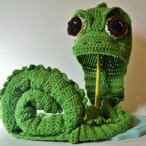 May include: Green crocheted snake hat and scarf set with large brown eyes. The scarf is coiled into a spiral shape.