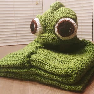 May include: Green crocheted blanket with a large crocheted chameleon head attached. The chameleon has large brown eyes with white pupils.
