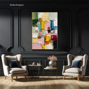May include: Abstract oil painting with a gold frame. The artwork features a colorful composition of rectangular shapes in shades of yellow, pink, blue, green, and red. The painting hangs on a dark gray wall above two armchairs.