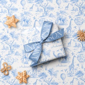 May include: A square gift box wrapped in white paper with a blue toile pattern featuring giraffes, elephants, and hot air balloons. A blue satin ribbon printed with the name Anderson is tied in a bow around the box. Three woven straw stars are nearby.
