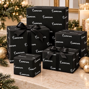 May include: Four rectangular gift boxes wrapped in matte black paper featuring the name Cameron printed in a white cursive script. Two of the boxes are tied with wide black satin ribbons tied into bows, while the other two remain untied.