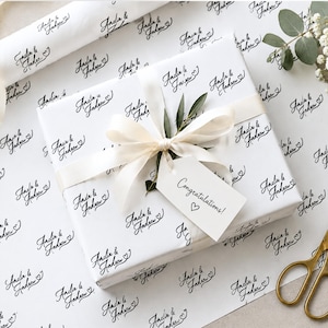 May include: A square gift box wrapped in white paper featuring a repeating black script pattern that reads Amelia & Andrew with a small heart. The box is tied with a cream ribbon, a green leaf sprig, and a white tag labeled Congratulations! with a heart.