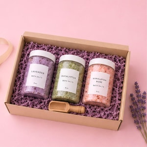 May include: A cardboard gift box containing three clear plastic jars of 8 oz bath salts labeled Lavender, Eucalyptus, and Himalayan Rose, each with a white lid. A small wooden scoop rests in the purple shredded paper filler next to the jars.