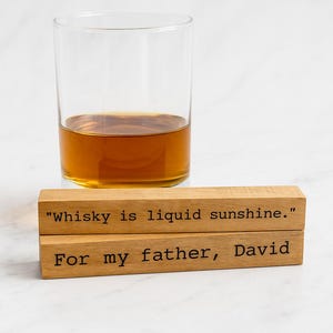 Puede incluir: Un vaso transparente con líquido ámbar, probablemente whisky, se encuentra sobre dos bloques de madera. El bloque superior dice: "whisky is liquid sunshine". El bloque inferior dice: "Para mi padre, David".