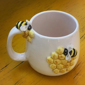 May include: White ceramic mug with a bee and honeycomb design. The mug features a curved handle and is decorated with a 3D bee and honeycomb pattern in yellow, black, and white. The mug is set against a yellow background.