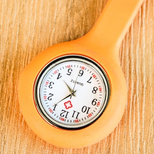 May include: Close-up of an orange quartz nurse fob watch. The watch face is white with black numerals and hands, and a red cross symbol. The watch has a pin for attaching to clothing. The watch is on a wooden surface.