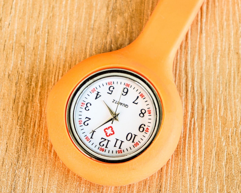 May include: Close-up of an orange quartz nurse fob watch. The watch face is white with black numerals and hands, and a red cross symbol. The watch has a pin for attaching to clothing. The watch is on a wooden surface.