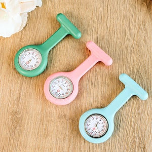 May include: Three colorful fob watches in green, pink, and light blue. Each watch has a white clock face with black numbers and red hands. The watches are designed with a T-shaped top for easy attachment.