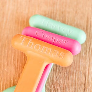 May include: Three colorful, personalized bottle openers. The orange opener reads "Thomas", the pink one says "Cooper", and the green one says "Harvey". The openers are arranged diagonally against a wooden background.