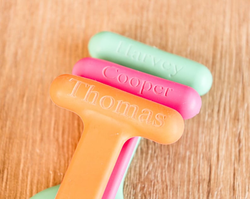 May include: Three colorful, personalized bottle openers. The orange opener reads "Thomas", the pink one says "Cooper", and the green one says "Harvey". The openers are arranged diagonally against a wooden background.