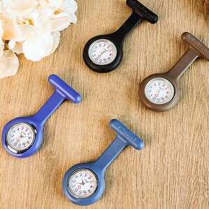 May include: Four nurse fob watches in black, brown, and blue. Each watch has a white face with black numbers and hands. The watches are on a wooden surface, with a few white flowers in the background. The watch faces are approximately 1 inch in diameter.