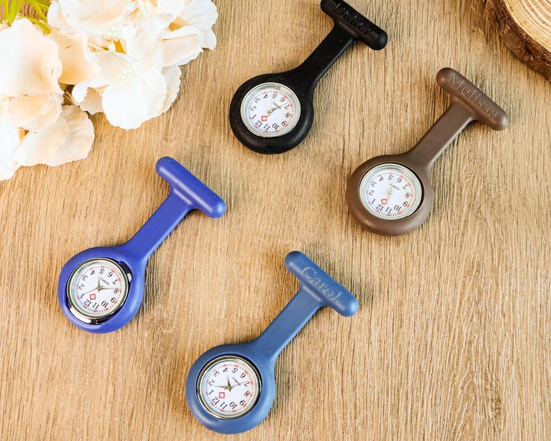 May include: Four nurse fob watches in black, brown, and blue. Each watch has a white face with black numbers and hands. The watches are on a wooden surface, with a few white flowers in the background. The watch faces are approximately 1 inch in diameter.