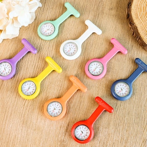 May include: A selection of colourful nurse fob watches in shades of purple, green, yellow, orange, pink, blue, white, and red. Each watch features a white clock face with black numerals and hands. The watches are displayed on a wooden surface.