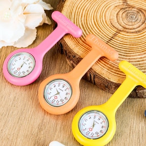 May include: Three colorful fob watches in pink, orange, and yellow. Each watch has a white face with black numerals and hands. The pink watch has "Cooper" printed on the handle, the orange watch has "Thomas", and the yellow watch has "Harvey".