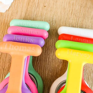 May include: A collection of colorful, personalized name badge holders. The holders are in various colors, including orange, yellow, green, pink, purple, red, and white. Each holder has a name printed on it in a contrasting color. The scissors are visible.