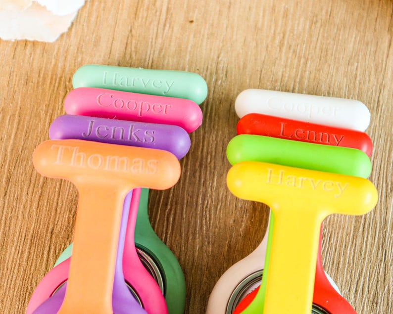 May include: A collection of colorful, personalized name badge holders. The holders are in various colors, including orange, yellow, green, pink, purple, red, and white. Each holder has a name printed on it in a contrasting color. The scissors are visible.
