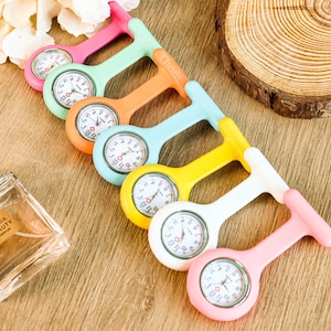 May include: A collection of colorful nurse fob watches in pink, green, orange, blue, yellow, and white. Each watch has a round face with black numbers and hands. The watches are arranged on a wooden surface.