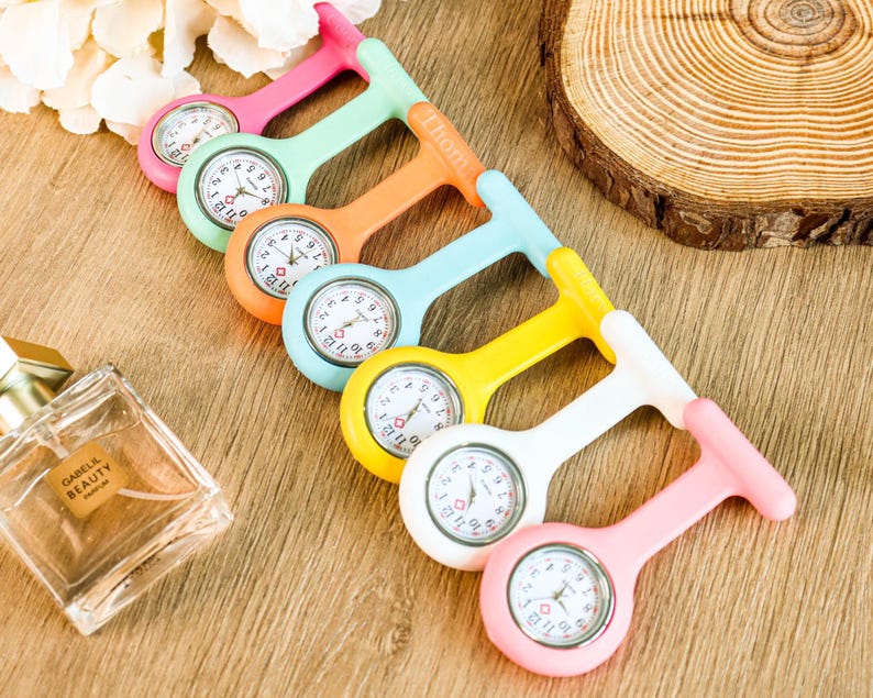 May include: A collection of colorful nurse fob watches in pink, green, orange, blue, yellow, and white. Each watch has a round face with black numbers and hands. The watches are arranged on a wooden surface.