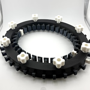 May include: A circular black 3D printed frame featuring a series of evenly spaced dark gray clips along the inner and outer edges, secured by multiple white four-pointed star-shaped knobs positioned around the top surface.