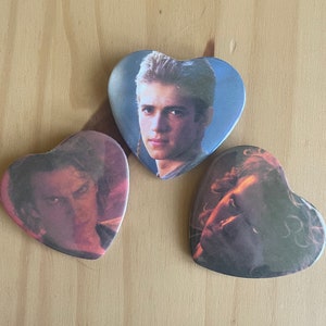 May include: Three heart-shaped buttons featuring images of a person. The central button displays a portrait of a person with light hair. The other two buttons show different images of the same person. The buttons are set against a light brown wooden surface.