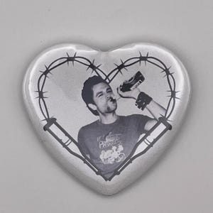 May include: Heart-shaped button with a black and white image of a person drinking from a bottle. The image is surrounded by a barbed wire design. The person is wearing a t-shirt with a graphic.