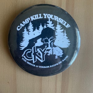 May include: Black button with white text and graphics. The text reads "CAMP KILL YOURSELF" above a silhouette of a person with a gun, trees, and a tent. The text "CKY" and "THE SHOCK & TERROR KEEPS ME SANE" are also present.