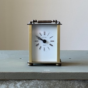 May include: A rectangular, gold-toned clock with a white face and black Roman numerals. The clock has a handle on top and sits on a gray surface. The clock face reads "SOTIMA Quartz".