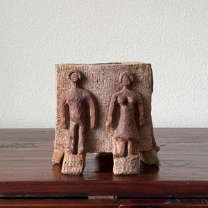 May include: A square-shaped terracotta planter featuring a rough, textured surface. Two sculpted human figures are attached to the front, each standing on a small base. The clay has a natural, earthy brown tone with visible markings and indentations.