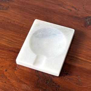 May include: A square white marble ashtray featuring a smooth polished finish, a central circular basin for holding items, and a single rectangular notch carved into one side for resting objects. The stone displays subtle grey veining throughout.