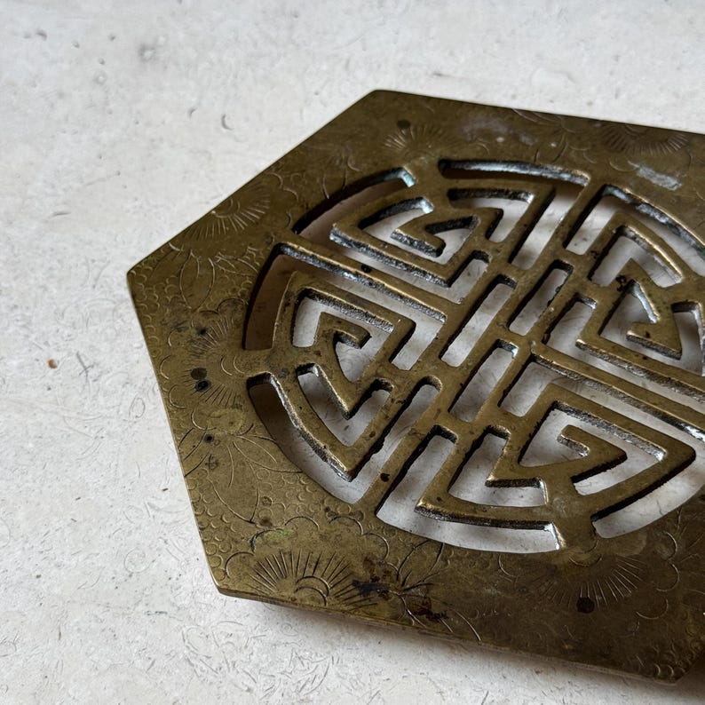 May include: A hexagonal brass coaster with a decorative, intricate design. The coaster features a circular cutout with a geometric pattern and a floral design around the perimeter.