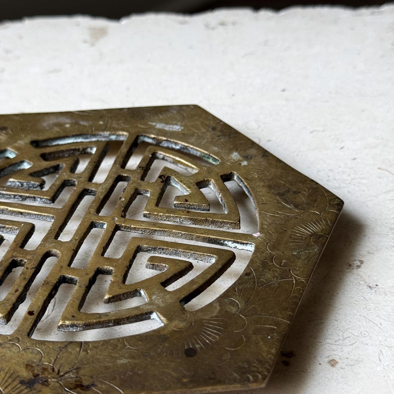 May include: A brass coaster with a hexagonal shape and an intricate, circular, cut-out design. The design features a repeating pattern of squares and triangles.