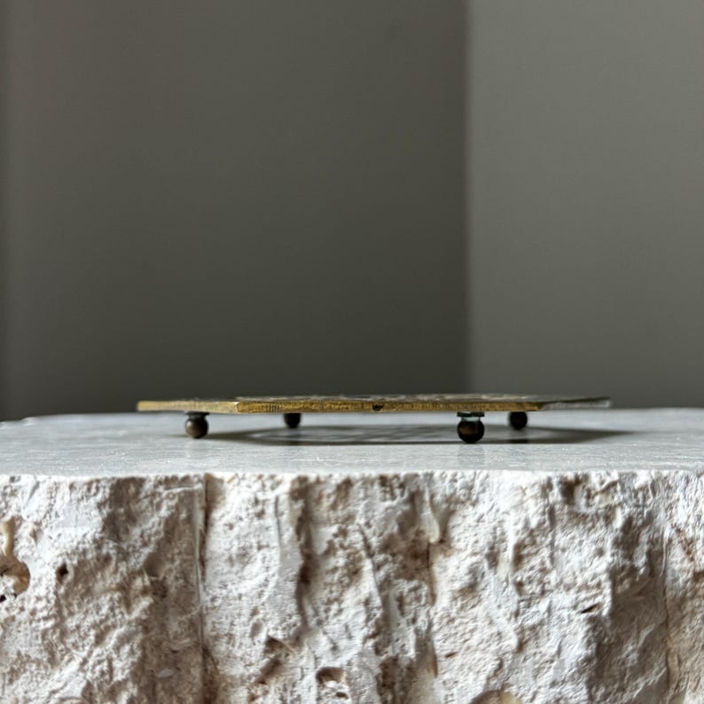 May include: A brass trivet with a geometric design and four small feet. The trivet is sitting on a white surface.