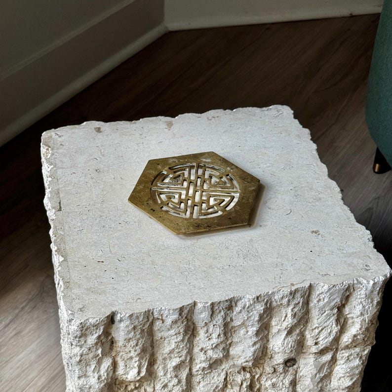 May include: A white stone side table with a carved, textured surface. The table has a brass hexagonal coaster with a decorative design.