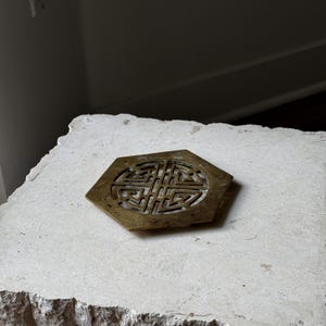May include: A brass coaster with a hexagonal shape and a decorative, intricate design in the center. The coaster is sitting on a light beige surface.