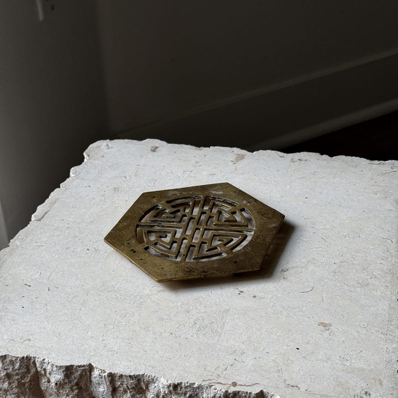 May include: A brass coaster with a hexagonal shape and a decorative, intricate design in the center. The coaster is sitting on a light beige surface.