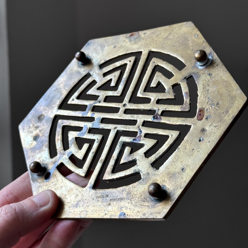 May include: A brass hexagonal coaster with a cut-out design of a Chinese knot. The coaster has a "Made in Hong Kong" inscription on the bottom.