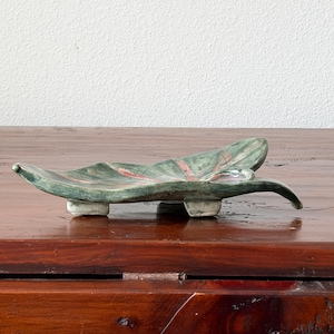 May include: A ceramic dish shaped like a leaf with a green and brown mottled glaze. The dish features raised veins in a reddish-brown hue and is supported by three small, light-colored feet, giving it a slightly elevated and organic appearance.