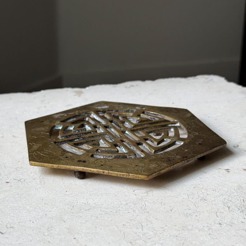 May include: A hexagonal brass coaster with an intricate, cut-out design. The coaster has three small feet on the bottom.
