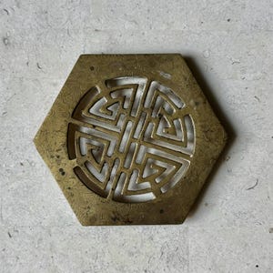 May include: A hexagonal brass coaster with a decorative, cut-out design in the center. The design is a circular pattern of interlocking squares.
